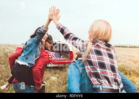 Vista posteriore di giovani donne piggybacking su fidanzati e dando alta cinque nel campo dei fiori Foto Stock