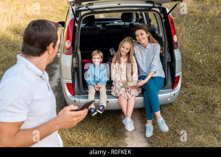 L'uomo utilizza lo smartphone mentre la sua famiglia seduta nel baule auto nel campo Foto Stock