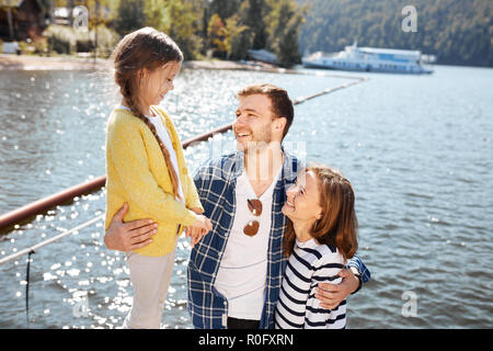 La famiglia felice di trascorrere del tempo insieme al di fuori del vicino lago. I genitori a giocare con la figlia abbracciando e divertirsi Foto Stock