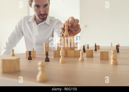 Grave giovane imprenditore re di posizionamento pezzo degli scacchi sulla sommità della piramide fatta di cubi di legno su una scrivania in ufficio con altri pezzi di scacchi posto su di esso. Foto Stock