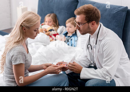 Pediatra dando pillole alla madre dei bambini malati che giace nel letto sullo sfondo Foto Stock