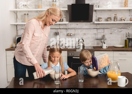 Madre versando il latte in cereali per gli adorabili bambini piccoli durante la prima colazione Foto Stock