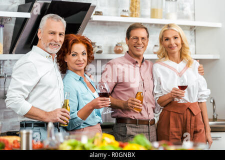 Felice coppia amici tenendo i bicchieri di vino e le bottiglie di birra, sorridente in telecamera in cucina Foto Stock