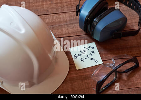 La sicurezza prima di tutto nota uno giallo pezzo di carta bloccata su sfondo di legno con una gamma completa di attrezzature per la protezione personale: casco, occhiali safet e orecchio p Foto Stock