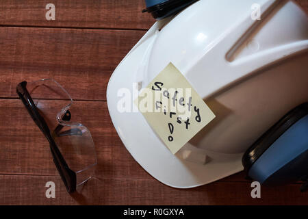 La sicurezza prima di tutto nota uno giallo pezzo di carta bloccata su sfondo di legno con una gamma completa di attrezzature per la protezione personale: casco, occhiali safet e orecchio p Foto Stock