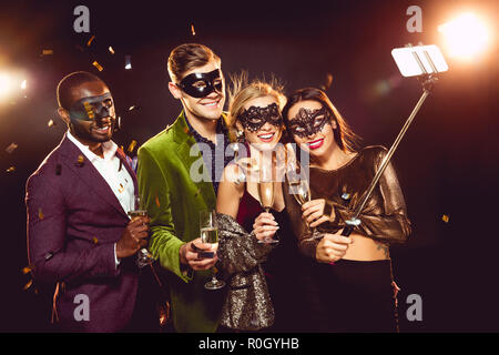 Glamour amici multiculturale in maschere di carnevale tenendo selfie sullo smartphone Foto Stock