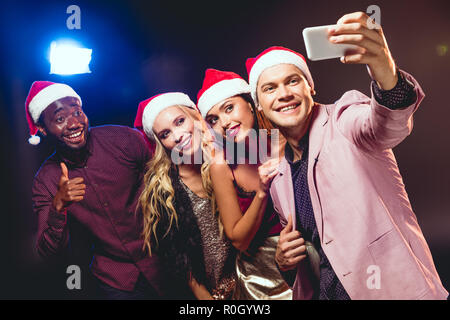 Glamour amici multiculturale in cappelli di Babbo Natale tenendo selfie sullo smartphone Foto Stock