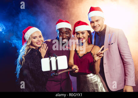 Happy amici multiculturale in cappelli di Babbo Natale con il segno della vittoria prendendo selfie sullo smartphone Foto Stock