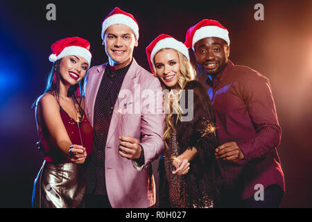 Allegro amici multiculturale in cappelli di Babbo Natale per celebrare il nuovo anno con luci di bengala Foto Stock