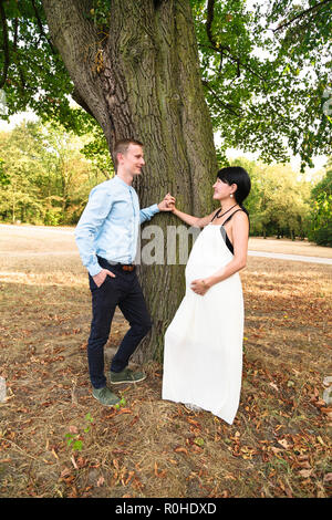 Famiglia internazionale con la donna incinta in piedi vicino a tree in posizione di parcheggio Foto Stock