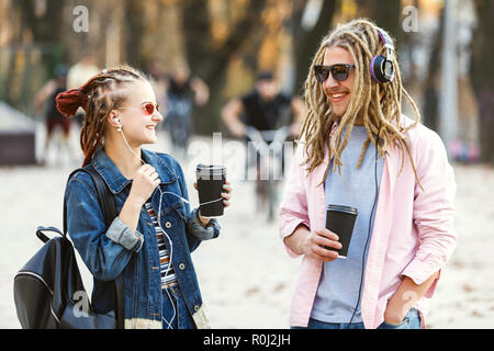 Due hipster migliori amici con i timori di indossare le cuffie e occhiali camminare insieme, di ridere e di bere il caffè in strada Foto Stock