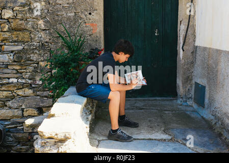 Ragazzo legge un libro a fumetti seduto di fronte a una casa di villaggio in Italia Foto Stock