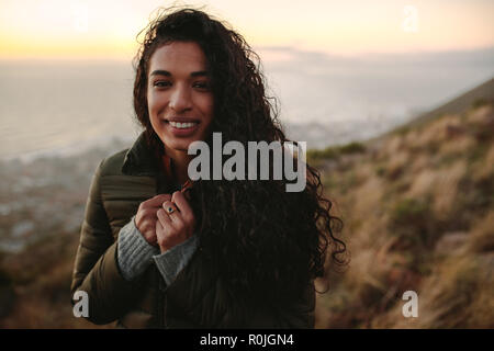 Ritratto di giovane donna africana con capelli ricci di indossare abiti caldi in piedi sulla scogliera. Femmina in natura sul giorno d'inverno. Foto Stock