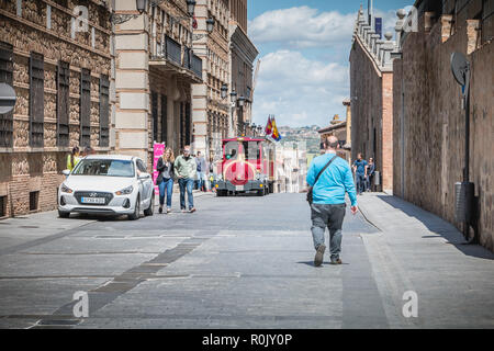 Toledo, Spagna - 28 Aprile 2018: turistici passeggiando in una strada del centro storico accanto a un piccolo turista trenino rosso su una giornata di primavera Foto Stock