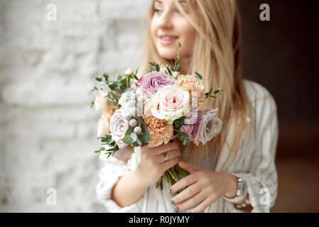 Close-up giovane donna caucasica holding bouquet di fiori, guarda attraverso la finestra con il sorriso sulla faccia in piedi nella stanza di luce in ambienti interni Foto Stock