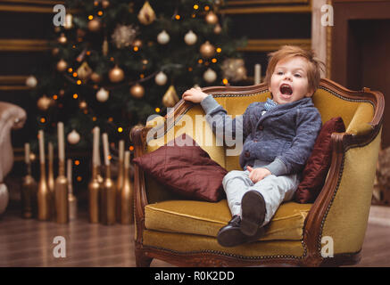 Un elegante bambino si siede in una sedia antica in previsione della Santa. La Vigilia di Capodanno e Natale. Una camera in stile classico. Un sacco di luci e di candela Foto Stock