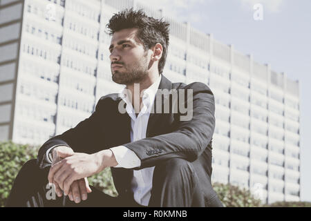 Fiducioso giovane uomo di successo seduta nella città. Dietro di lui un ufficio moderno edificio. Capelli alla moda, una camicia bianca e giacca. Foto Stock