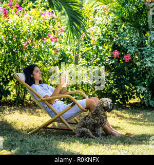 Giovane donna seduta in sedia a sdraio e la lettura di un libro, cane, giardino tropicale, Guadalupa, French West Indies, Foto Stock