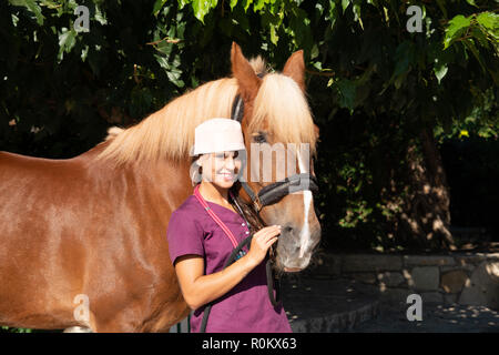 Sorridente giovane femmina medico veterinario ritratto con cavallo marrone all'aperto. Assistenza sanitaria equestre concetto medico. Foto Stock