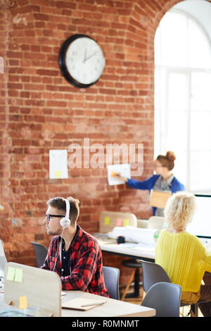 Giovani manager in cuffie e due suoi colleghi seduti dai loro luoghi di lavoro e di lavoro in ufficio Foto Stock