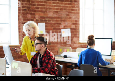 Matura femmina fiducioso puntando sullo schermo del computer mentre spiegando il suo giovane collega alcuni momenti di organizzazione Foto Stock