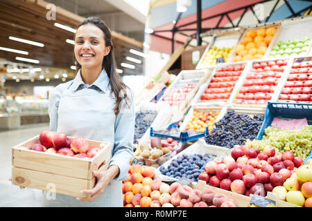 Allegro eccitato giovane femmina rivenditore di generi alimentari in grembiule in piedi contro prodotti alimentari e di stallo della scatola di contenimento di mele, ella la vendita di frutta e verdura fresca Foto Stock