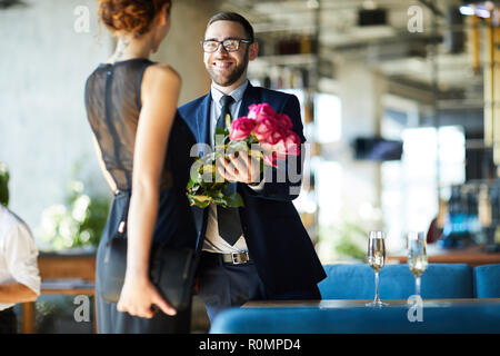 Felice giovane uomo dando mazzo di rose rosa alla sua ragazza durante la data romantica nel ristorante Foto Stock