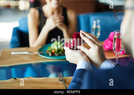 Anello diamante in rossa piccola scatola di velluto detenute da giovane uomo rendendo proposta alla sua ragazza in ristorante Foto Stock