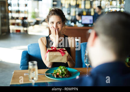 Giovane donna entusiasta della scatola di contenimento con inaspettato regalo dal suo fidanzato durante il pranzo in ristorante Foto Stock