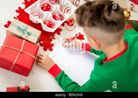 Carino il ragazzo che indossa il natale pigiami scrivere lettera a Babbo su livingroom piano. Vista aerea di un giovane ragazzo di scrivere il suo natale wishlist. Foto Stock