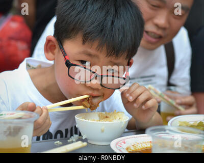 Little Chinese Boy con ingegnosi occhiali mangia riso al vapore e carne di maiale con bacchette di legno. Foto Stock