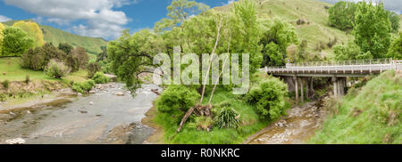 Un ponte di cemento con ringhiere in bianco che attraversa un fiume con verdi colline sullo sfondo sotto il cielo blu con nuvole bianche Foto Stock