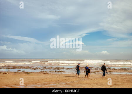 Una famiglia non identificabili camminando lungo una spiaggia disseminato dopo una tempesta con il bianco delle nuvole, cielo azzurro, sabbia marrone e molte onde che si infrangono sulla riva Foto Stock