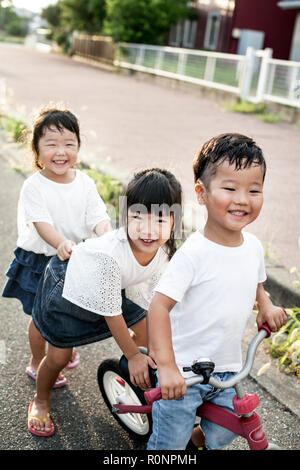 Ritratto di due ragazze giapponesi e ragazzo giocando sulla strada con una bicicletta, sorridente alla fotocamera. Foto Stock