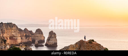 Escursionista guarda oltre il tramonto sul mare costa rocciosa di arenaria, formazioni rocciose nel mare, Algarve, Lagos, Portogallo Foto Stock