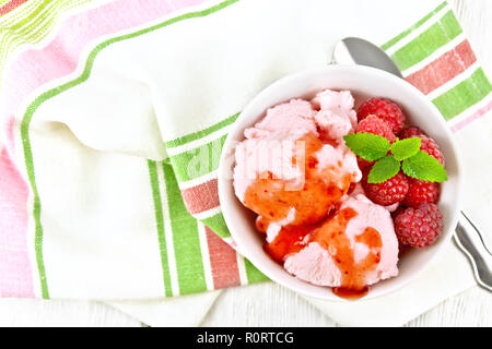 Ice Cream crimson con bacche di lampone, e sciroppo di menta in vaso bianco, un cucchiaio su asciugamano contro luce pannello di legno sulla parte superiore Foto Stock