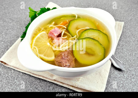 Minestra con zucchine, carni bovine, prosciutto, limone e tagliatelle in una ciotola su un tovagliolo, il prezzemolo e l'aneto su un tavolo di granito sullo sfondo Foto Stock