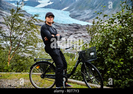 Giovane uomo in abiti neri sulla bicicletta, con cappuccio nero, ghiacciaio Svartisen sullo sfondo Foto Stock