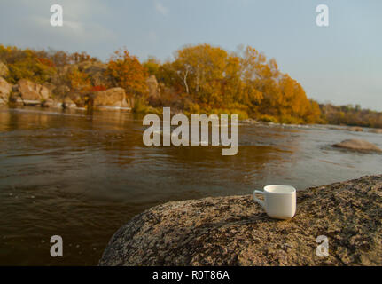 Una tazza di caffè si erge su una pietra sulla banca del fiume; l'autunno è di colore giallo brillante e alberi di arancio circondato da Foto Stock