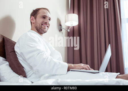 Sorridente uomo piacevole indossare accappatoio bianco trascorrere la mattina in hotel Foto Stock