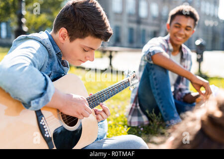 Felice adolescenti seduti su erba e suonare la chitarra acustica Foto Stock