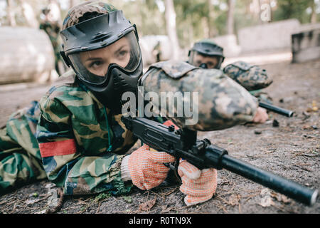 Paintball femmina player nella maschera goggle e camouflage con pistola per marcatore strisciando sul terreno all'aperto Foto Stock