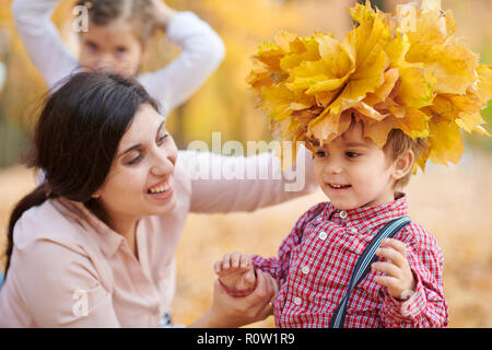 Mamma mette il giallo caduta foglie sul figlio di testa. La famiglia felice è in autunno il parco della città. Bambini e genitori. Essi pongono, sorridente, giocare e divertirsi. Foto Stock