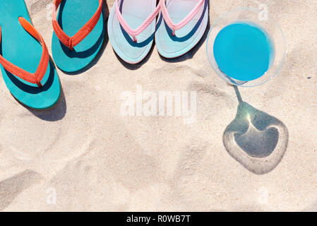 Viaggi e summer party. Pantofole colorate sulla spiaggia con bevanda blu su bianco sabbia. Felici vacanze estive. Foto per aggiungere messaggio di testo. Sfondo f Foto Stock