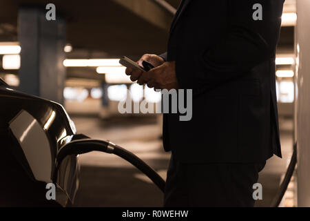Imprenditore utilizzando il telefono cellulare mentre si ricarica auto elettrica Foto Stock