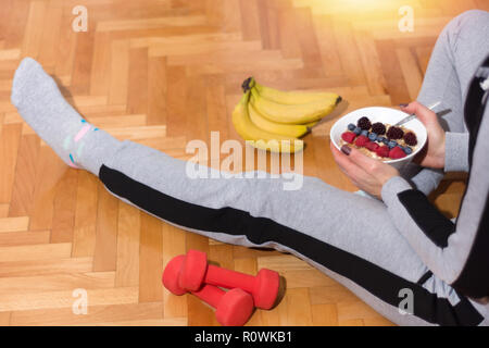 Donna dopo esercizi mangiare mangiare avena porridge in ciotola decorata con more, lamponi e mirtilli. Ragazza seduta sul pavimento con il manubrio Foto Stock