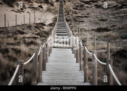 In legno e fatiscente weathered boardwalk con fune di ringhiere che conducono verso la collina sopra erboso dune di sabbia. La profondità di campo di una sfocatura Foto Stock