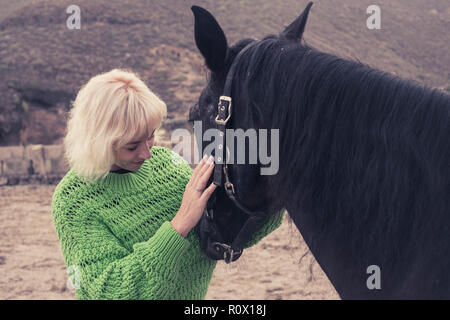 Allegro giovane donna capelli biondi con la sua incantevole scuro nero cavallo bellissimo. Di amore e di affetto per gli animali in outdoor le attività per il tempo libero. Wild e natur Foto Stock