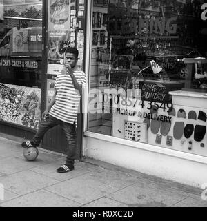 Ragazzo con il calcio sotto i suoi piedi, drink in mano,sulla strada al di fuori di un lavaggio a secco Negozio,Southwark broadway London REGNO UNITO Foto Stock