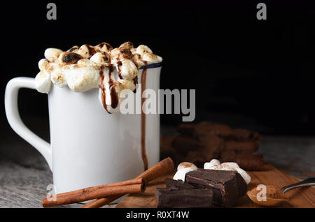 Primo piano di una tazza di cioccolata calda con marshmallows tostato con i cookie, aggiungete pezzetti di cioccolato, bastoncini di cannella, contro uno sfondo scuro. Foto Stock